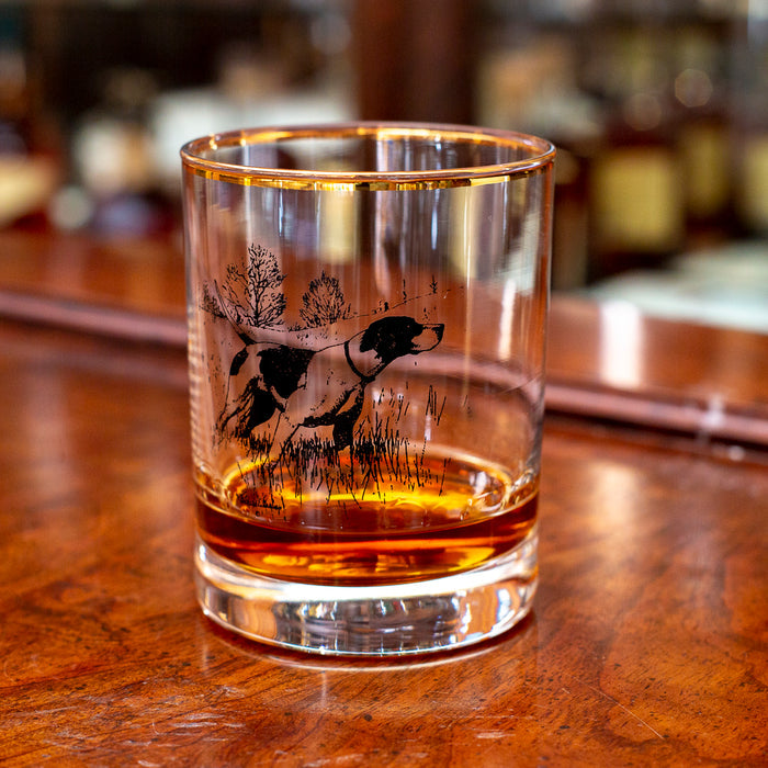 14 oz double old fashioned glass with a gold rim and black etched sporting dog artwork on a wooden bar top.