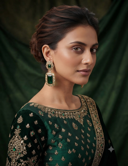 A woman wearing a dark green embroidered traditional outfit with gold detailing and matching green rectangular drop earrings, posed against a dark green draped backdrop.