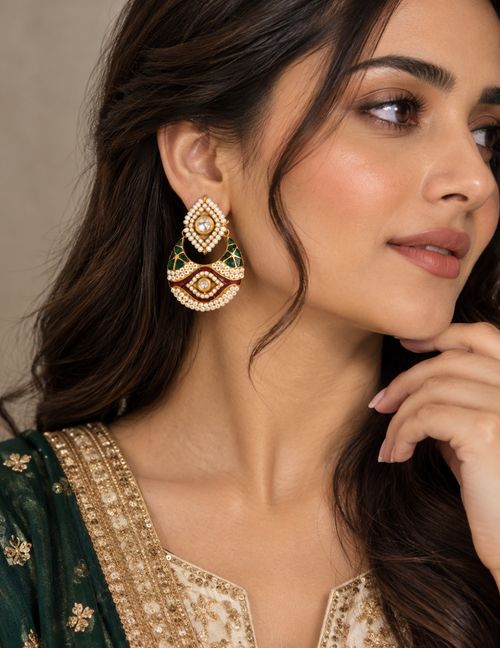 A close-up of a woman wearing a large, ornate gold-toned traditional Indian jhumka earring embellished with green, red, and white stones. She is wearing a dark green embroidered kurta.