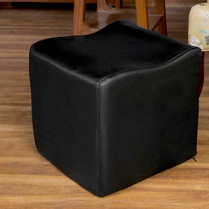Black cube-shaped ottoman on a wooden floor with a floral-patterned chair and wooden shelf in the background.