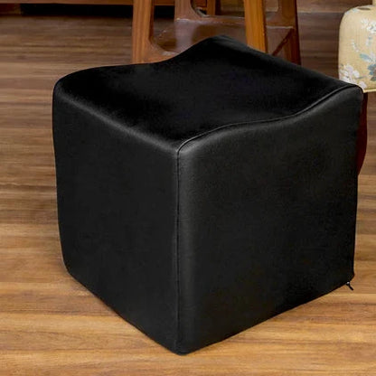Black cube-shaped ottoman on a wooden floor with a floral-patterned chair and wooden shelf in the background.