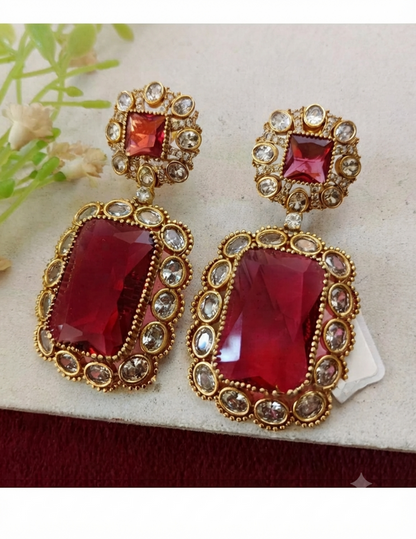A pair of gold-plated statement earrings with large rectangular red gemstones and smaller round clear stones, displayed on a light textured surface with a small white tag visible.